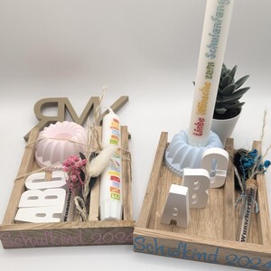 K&ouml;nnte beinhalten: Zwei Holztabletts mit der Aufschrift "Schulkid 2024" auf der Vorderseite. Jedes Tablett enth&auml;lt eine kleine Kuchenform, eine Kerze und Holzbuchstaben, die "ABC" buchstabieren. Ein Tablett ist in Rosa und Wei&szlig; dekoriert, w&auml;hrend das andere in Blau und Wei&szlig; dekoriert ist.