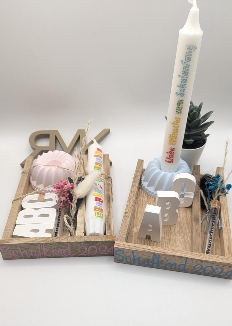 K&ouml;nnte beinhalten: Zwei Holztabletts mit der Aufschrift "Schulkid 2024" auf der Vorderseite. Jedes Tablett enth&auml;lt eine kleine Kuchenform, eine Kerze und Holzbuchstaben, die "ABC" buchstabieren. Ein Tablett ist in Rosa und Wei&szlig; dekoriert, w&auml;hrend das andere in Blau und Wei&szlig; dekoriert ist.