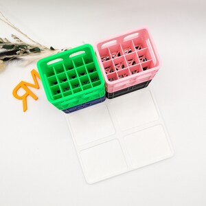 Op de afbeelding: Een stapel van drie kleurrijke plastic kratten, groen, roze en zwart, met een witte vierkante lade op de voorgrond. De kratten hebben een rasterpatroon aan de binnenkant. De achtergrond is wit.