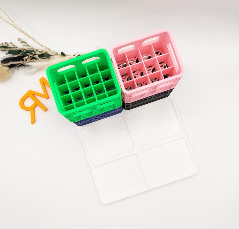 Op de afbeelding: Een stapel van drie kleurrijke plastic kratten, groen, roze en zwart, met een witte vierkante lade op de voorgrond. De kratten hebben een rasterpatroon aan de binnenkant. De achtergrond is wit.