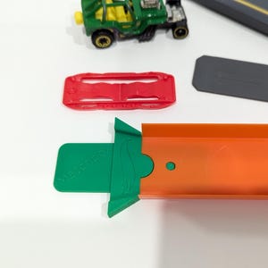 Peut inclure: Une voiture jouet verte avec une pi&egrave;ce de piste rouge et verte. La pi&egrave;ce de piste a un petit cercle vert sur le c&ocirc;t&eacute;. La voiture est sur une surface blanche.