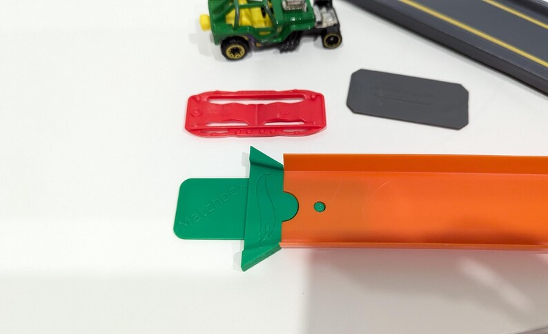 Peut inclure: Une voiture jouet verte avec une pi&egrave;ce de piste rouge et verte. La pi&egrave;ce de piste a un petit cercle vert sur le c&ocirc;t&eacute;. La voiture est sur une surface blanche.