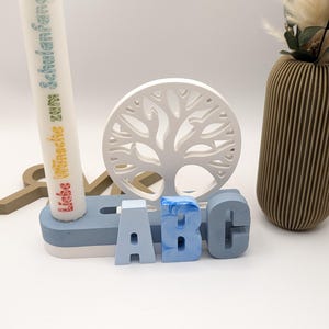 K&ouml;nnte beinhalten: Eine wei&szlig;e Kerze mit dem Text "Liebe W&uuml;nsche zum Schulanfang" in Regenbogenfarben, ein wei&szlig;es Holz-Baum-Ausschnitt und drei blaue Holzbuchstaben, die "ABC" auf einer blauen und wei&szlig;en Basis buchstabieren.