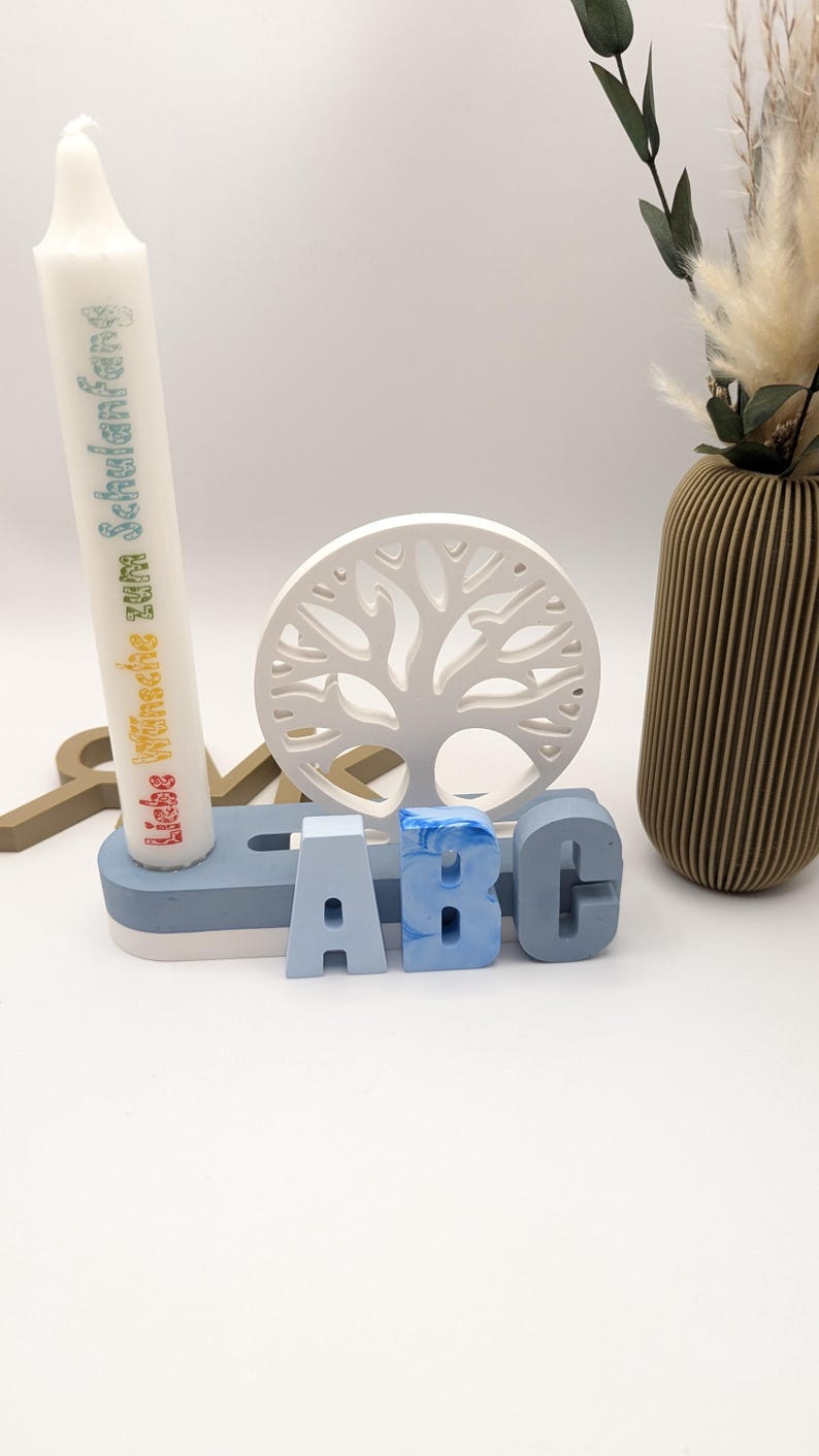 K&ouml;nnte beinhalten: Eine wei&szlig;e Kerze mit dem Text "Liebe W&uuml;nsche zum Schulanfang" in Regenbogenfarben, ein wei&szlig;es Holz-Baum-Ausschnitt und drei blaue Holzbuchstaben, die "ABC" auf einer blauen und wei&szlig;en Basis buchstabieren.