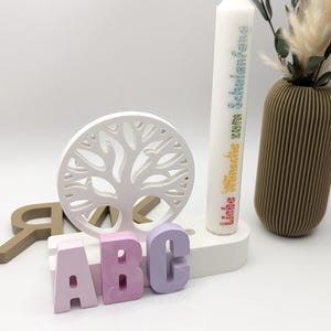 K&ouml;nnte beinhalten: Ein wei&szlig;er Holz-Lebensbaum mit einem Kerzenhalter und drei bunten Buchstaben, die "ABC" buchstabieren. Die Kerze ist wei&szlig; mit Regenbogentext, der "Liebe W&uuml;nsche zum Schulanfang f&uuml;r dich" lautet.