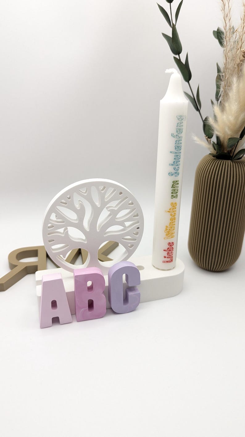 K&ouml;nnte beinhalten: Ein wei&szlig;er Holz-Lebensbaum mit einem Kerzenhalter und drei bunten Buchstaben, die "ABC" buchstabieren. Die Kerze ist wei&szlig; mit Regenbogentext, der "Liebe W&uuml;nsche zum Schulanfang f&uuml;r dich" lautet.