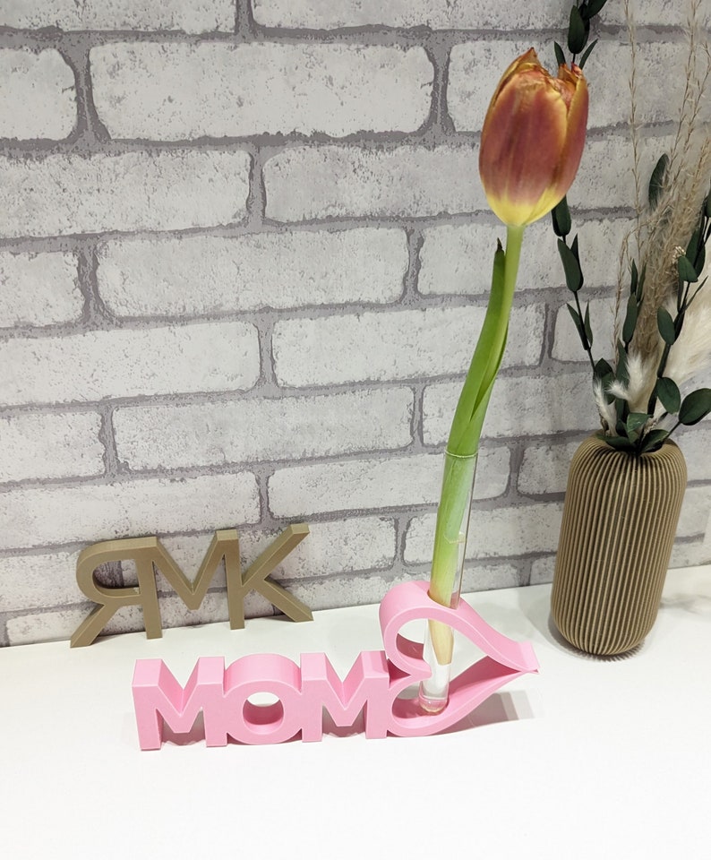 Pu&ograve; includere: Vaso in ceramica rosa a forma della parola "MOM" con un'apertura a forma di cuore per un solo fiore. Un solo tulipano arancione &egrave; nel vaso. Il vaso &egrave; su una superficie bianca con un muro di mattoni sullo sfondo.