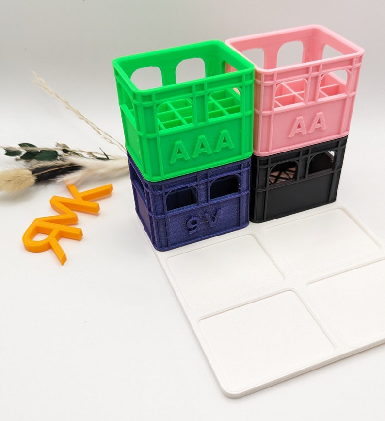 Op de afbeelding: Stapel kleurrijke miniatuurkratten in groen, blauw, roze en zwart, met de letters "AAA" en "9V" zichtbaar. Een witte tray met vier vierkante compartimenten staat op de voorgrond. Oranje letters spellen "NR".