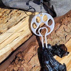 以下が含まれることがあります： 白いプラスチック製の犬の糞袋ディスペンサーで、足跡のデザインが施されています。ディスペンサーは、ジッパー付きのグレーのバッグに取り付けられています。黒いプラスチック製のバッグがディスペンサーからぶら下がっています。