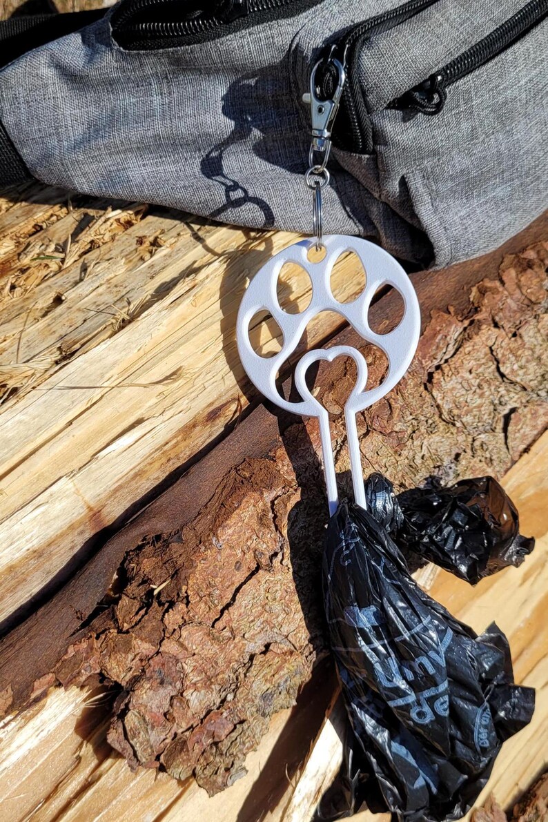 以下が含まれることがあります： 白いプラスチック製の犬の糞袋ディスペンサーで、足跡のデザインが施されています。ディスペンサーは、ジッパー付きのグレーのバッグに取り付けられています。黒いプラスチック製のバッグがディスペンサーからぶら下がっています。