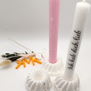 Peut inclure: Deux bougeoirs blancs avec des accents dor&eacute;s, chacun tenant une bougie blanche. La bougie de gauche est rose et porte le texte "f&uuml;r Dich". La bougie de droite est blanche et porte le texte "Ich hab dich lieb".