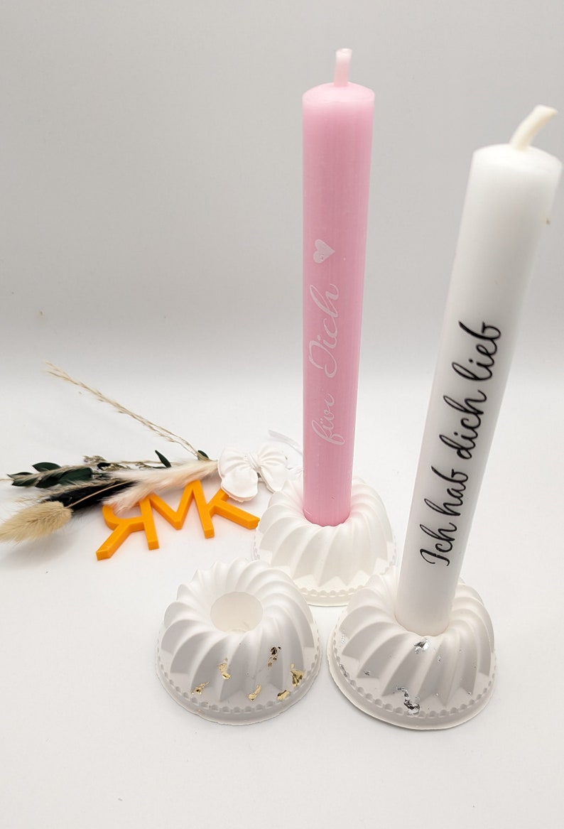Peut inclure: Deux bougeoirs blancs avec des accents dor&eacute;s, chacun tenant une bougie blanche. La bougie de gauche est rose et porte le texte "f&uuml;r Dich". La bougie de droite est blanche et porte le texte "Ich hab dich lieb".