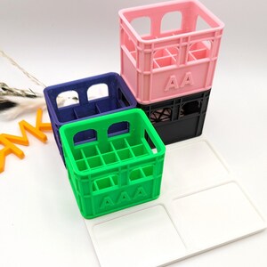Op de afbeelding: Een stapel kleurrijke miniatuurkratten in roze, blauw en groen, met een wit dienblad. Op de roze krat staat "AA". Op de groene krat staat "AAA". De kratten zijn op elkaar gestapeld.