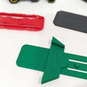 Peut inclure: Support de voiture Hot Wheels en plastique vert avec le texte "Racebox" imprim&eacute; dessus. Le support est con&ccedil;u pour maintenir une voiture Hot Wheels en position verticale.