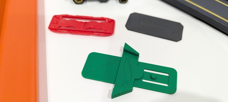 Peut inclure: Support de voiture Hot Wheels en plastique vert avec le texte "Racebox" imprim&eacute; dessus. Le support est con&ccedil;u pour maintenir une voiture Hot Wheels en position verticale.