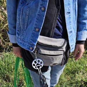 以下が含まれることがあります： 青いデニムジャケット、灰色のウエストバッグ、青いジーンズを着用した人。ウエストバッグには、足跡の付いた銀色の犬の糞袋ディスペンサーが取り付けられています。緑色のリードがディスペンサーからぶら下がっています。