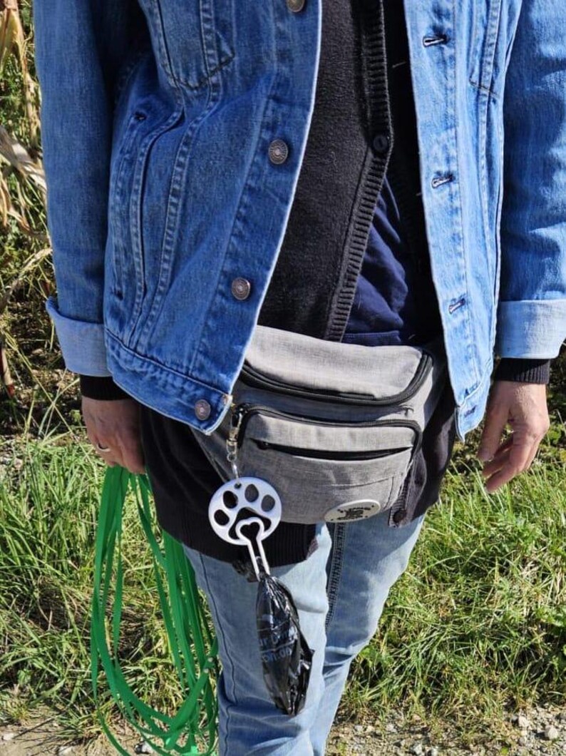 以下が含まれることがあります： 青いデニムジャケット、灰色のウエストバッグ、青いジーンズを着用した人。ウエストバッグには、足跡の付いた銀色の犬の糞袋ディスペンサーが取り付けられています。緑色のリードがディスペンサーからぶら下がっています。