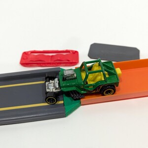 Peut inclure: Une voiture jouet verte avec une cage de s&eacute;curit&eacute; jaune roule sur une piste de jouet. La piste est compos&eacute;e de deux sections, une grise et une orange.
