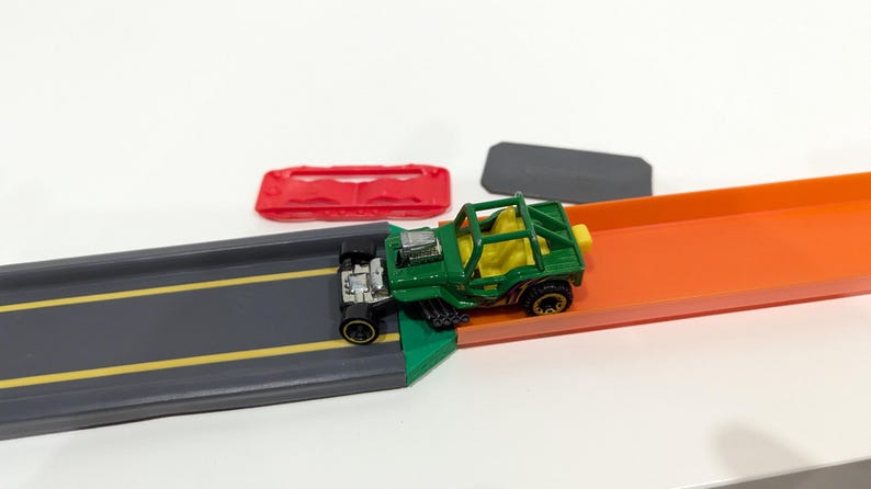 Peut inclure: Une voiture jouet verte avec une cage de s&eacute;curit&eacute; jaune roule sur une piste de jouet. La piste est compos&eacute;e de deux sections, une grise et une orange.