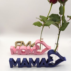 Pu&ograve; includere: Due vasi per fiori stampati in 3D a forma di cuore con la parola "MAMA" in rosa e blu. I vasi sono pieni di rose rosse.
