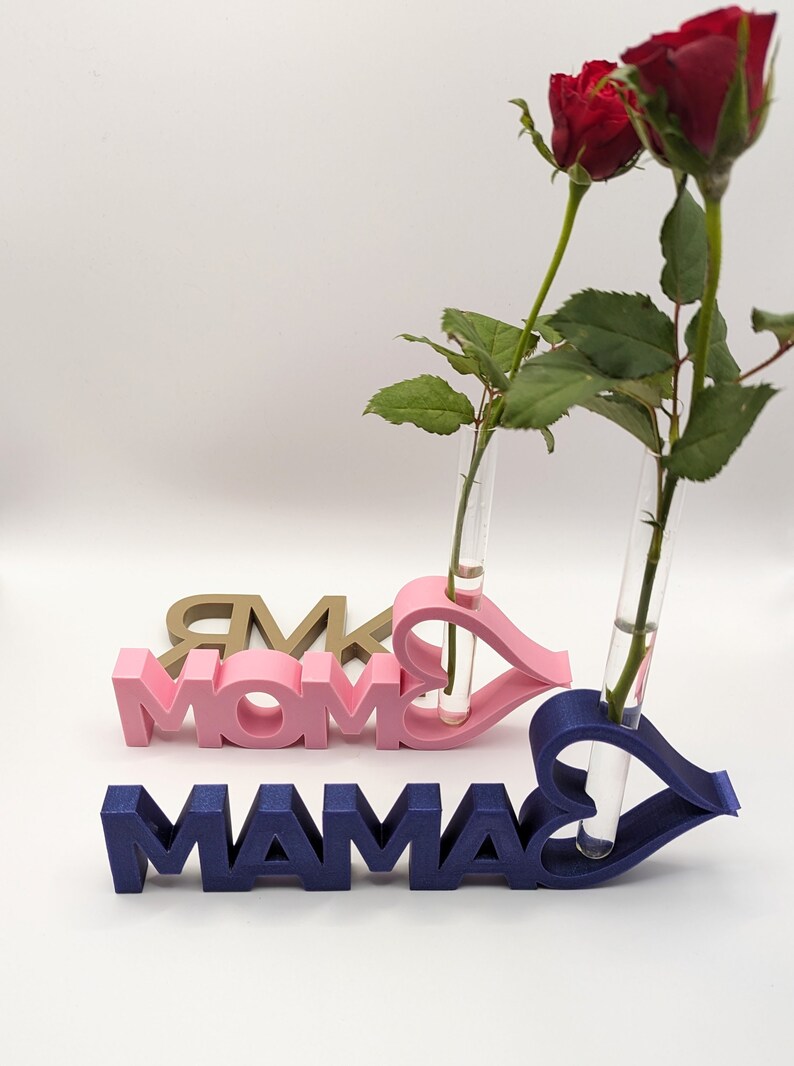 Pu&ograve; includere: Due vasi per fiori stampati in 3D a forma di cuore con la parola "MAMA" in rosa e blu. I vasi sono pieni di rose rosse.