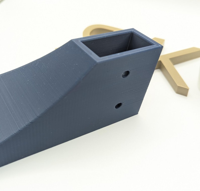 Puede incluir: Un soporte de esquina de pl&aacute;stico impreso en 3D de color azul oscuro con dos agujeros. El soporte tiene una abertura rectangular en la parte superior.