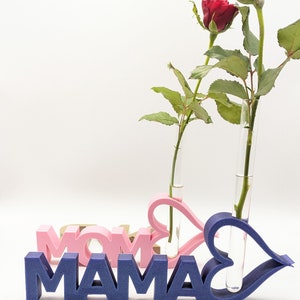 Pu&ograve; includere: Un cartello in legno blu e rosa che dice "MAMA" e "MOM" con due vasi di vetro a forma di cuore che contengono rose rosse.