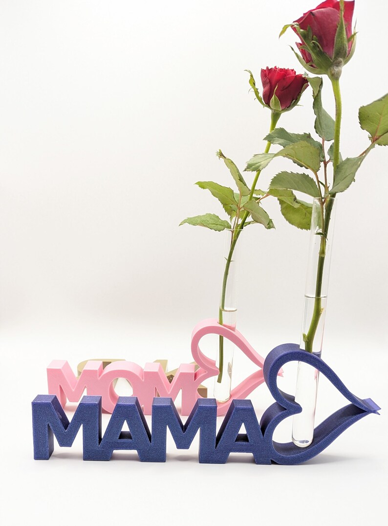 Pu&ograve; includere: Un cartello in legno blu e rosa che dice "MAMA" e "MOM" con due vasi di vetro a forma di cuore che contengono rose rosse.