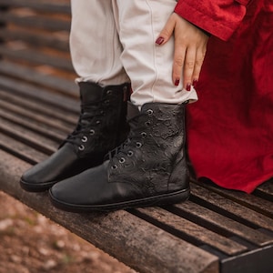 Könnte beinhalten: Schwarze Leder-Schnürstiefel mit geprägten Blumenmustern. Die Stiefel haben eine runde Spitze und einen kleinen Absatz. Die Stiefel werden mit hellen Hosen und einem roten Mantel getragen. Die Stiefel sitzen auf einer Holzbank.