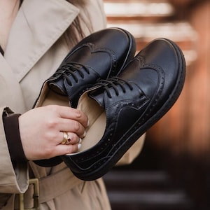 Puede incluir: Un par de zapatos Oxford de cuero negro con perforaciones decorativas y un dise&ntilde;o en forma de coraz&oacute;n en el lateral. Los zapatos tienen cordones negros y una suela gruesa. Los zapatos son sostenidos por una persona.