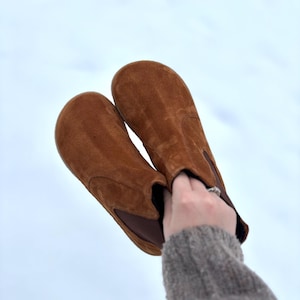 May include: A pair of brown suede ankle boots with a rounded toe and elastic side panels. The boots are held up against a light blue background, showcasing their warm, earthy tones and casual style.
