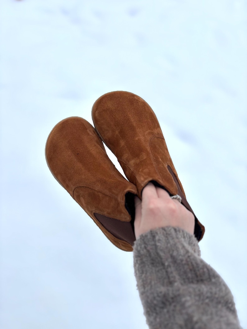 May include: A pair of brown suede ankle boots with a rounded toe and elastic side panels. The boots are held up against a light blue background, showcasing their warm, earthy tones and casual style.