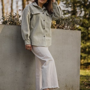 May include: A woman wearing a light green jacket, white pants, and white sneakers. She is standing next to a concrete wall and has her hand in her hair. She is wearing round sunglasses.