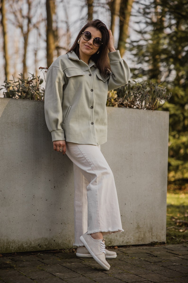 May include: A woman wearing a light green jacket, white pants, and white sneakers. She is standing next to a concrete wall and has her hand in her hair. She is wearing round sunglasses.