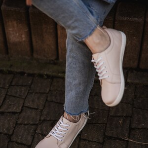May include: A pair of light pink leather sneakers with white laces. The sneakers are worn by a person wearing blue jeans.