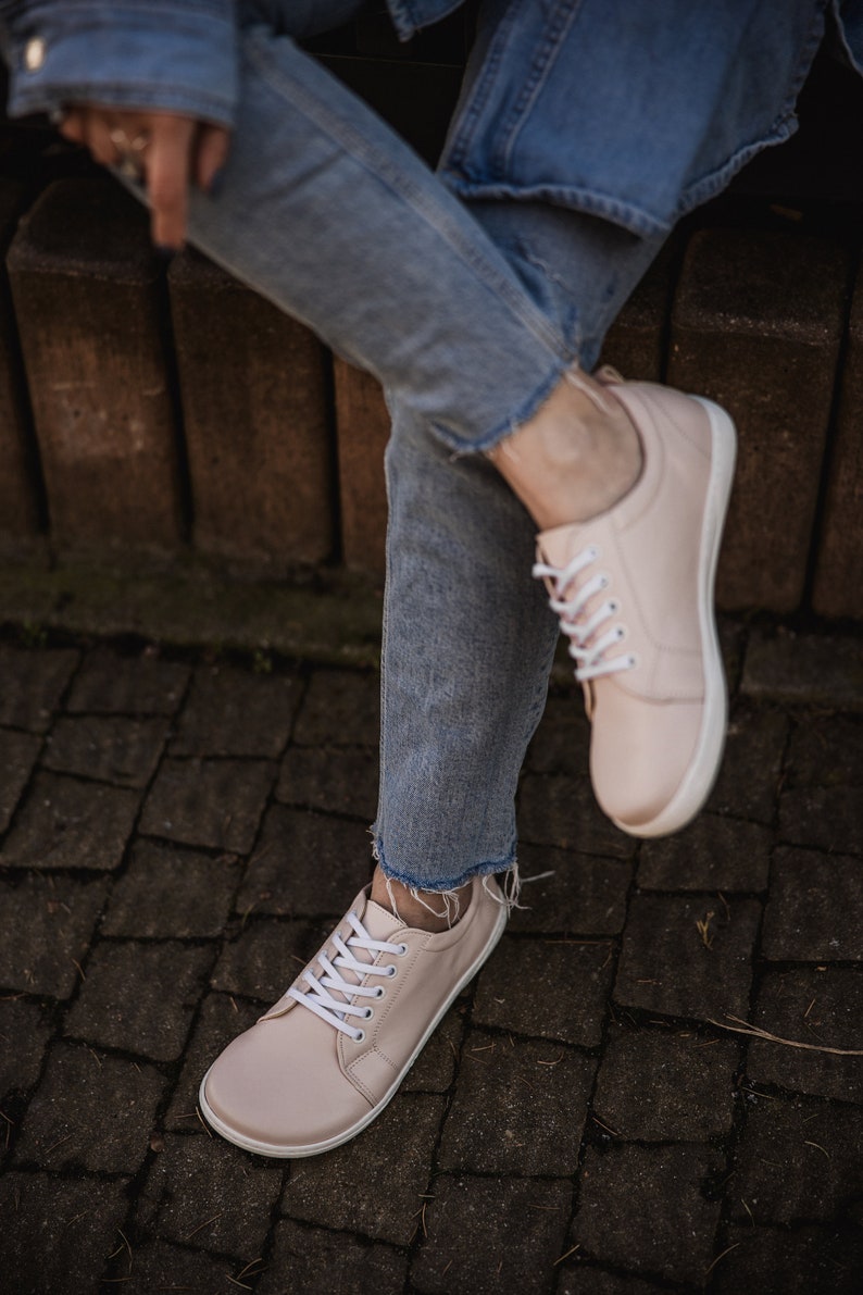 May include: A pair of light pink leather sneakers with white laces. The sneakers are worn by a person wearing blue jeans.