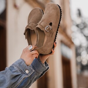 Peut inclure: Une paire de chaussures en daim marron clair avec une boucle de fermeture. Les chaussures ont une semelle en caoutchouc noir et sont tenues par les mains d'une personne.