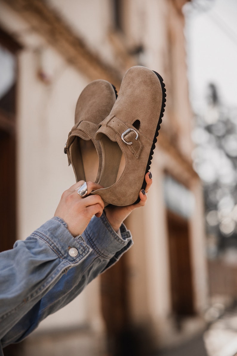 Peut inclure: Une paire de chaussures en daim marron clair avec une boucle de fermeture. Les chaussures ont une semelle en caoutchouc noir et sont tenues par les mains d'une personne.