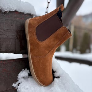 May include: A brown suede ankle boot with a dark brown elastic side panel. The boot has a light brown sole and is set against a snowy background. The boot is suspended from a chain.
