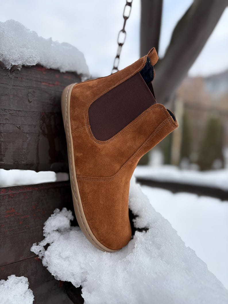 May include: A brown suede ankle boot with a dark brown elastic side panel. The boot has a light brown sole and is set against a snowy background. The boot is suspended from a chain.