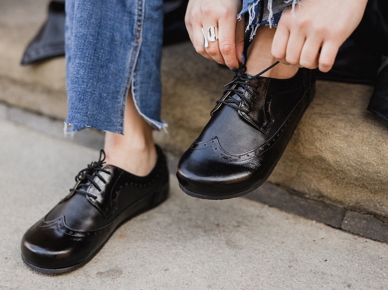 Pu&ograve; includere: Scarpe brogue in pelle nera con finitura lucida. Le scarpe presentano decorazioni traforate e chiusura con lacci. L'immagine mostra le scarpe che vengono allacciate. Le scarpe sono abbinate a jeans blu.