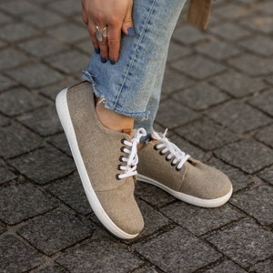 May include: A pair of light brown canvas trainers with white laces and white soles. The trainers are being worn by someone wearing blue jeans.