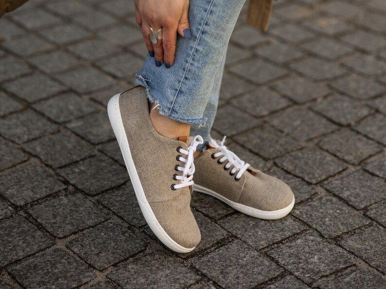 May include: A pair of light brown canvas trainers with white laces and white soles. The trainers are being worn by someone wearing blue jeans.