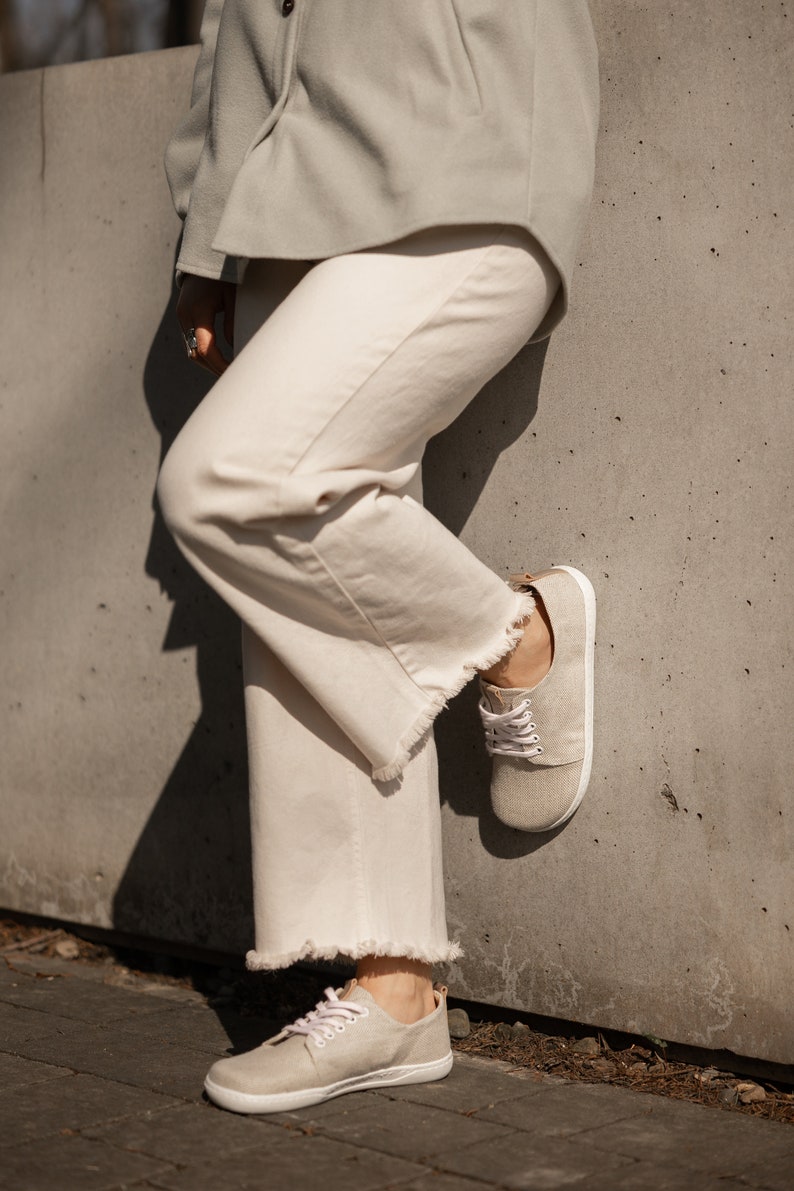 May include: A pair of light beige canvas shoes with white laces. The shoes have a rounded toe and a flat sole. The shoes are worn by a person wearing white pants and a light green jacket.
