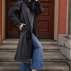 Pu&ograve; includere: Una persona indossa un trench coat lungo in pelle nera, jeans blu e scarpe nere con occhiali da sole rotondi. Il cappotto ha una cintura. I jeans sono svasati con spacco sull'orlo. Le scarpe sono stringate.
