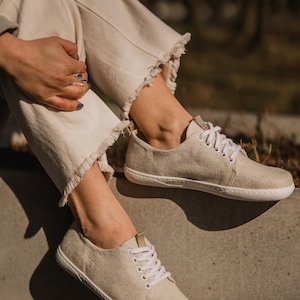 May include: A pair of beige canvas low-top sneakers with white laces. The shoes are worn by a person sitting on a concrete surface.