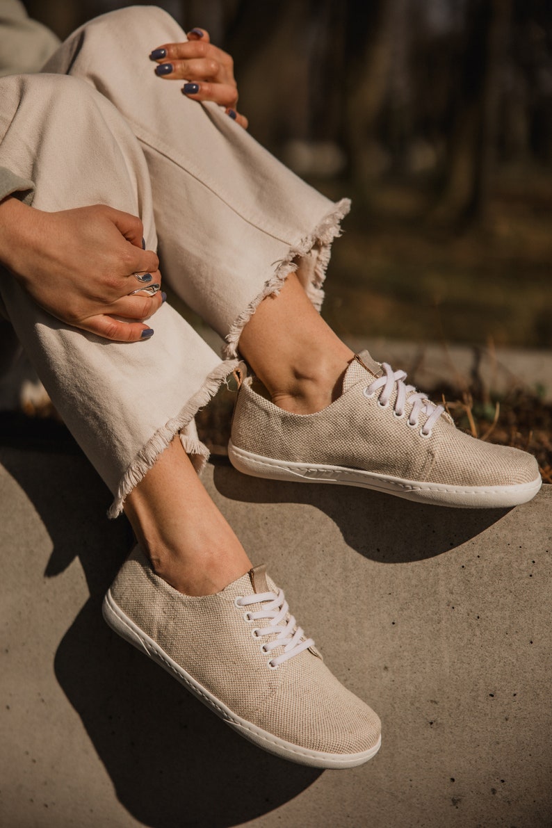 May include: A pair of beige canvas low-top sneakers with white laces. The shoes are worn by a person sitting on a concrete surface.