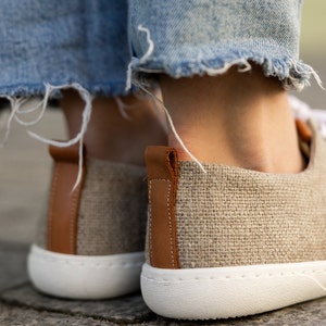 May include: A pair of beige canvas shoes with brown leather accents. The shoes have white soles and are made of a textured fabric.