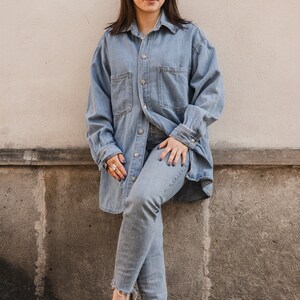 Peut inclure: Une femme portant une chemise en jean bleu clair et un jean, avec une paire de sabots en daim marron. Elle est debout devant un mur blanc.