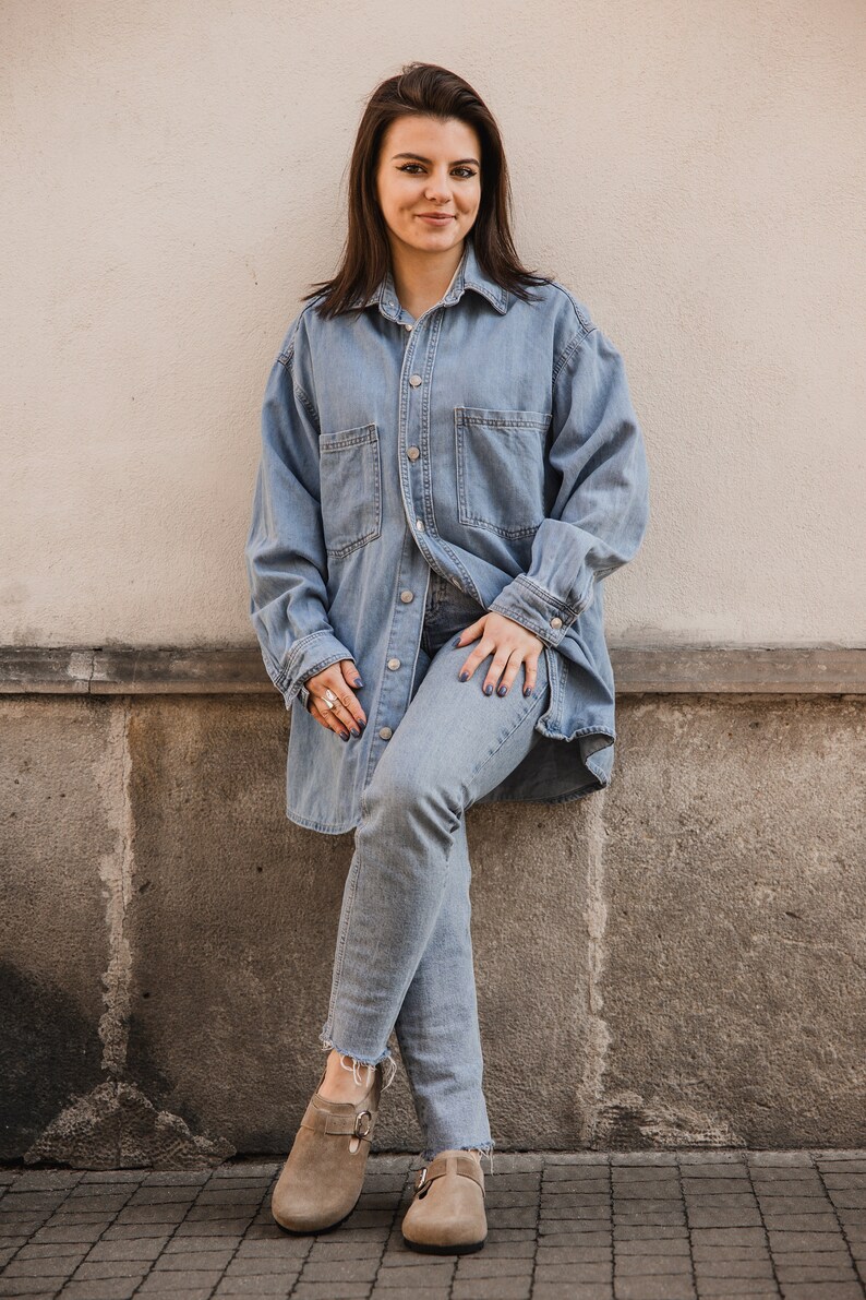 Peut inclure: Une femme portant une chemise en jean bleu clair et un jean, avec une paire de sabots en daim marron. Elle est debout devant un mur blanc.